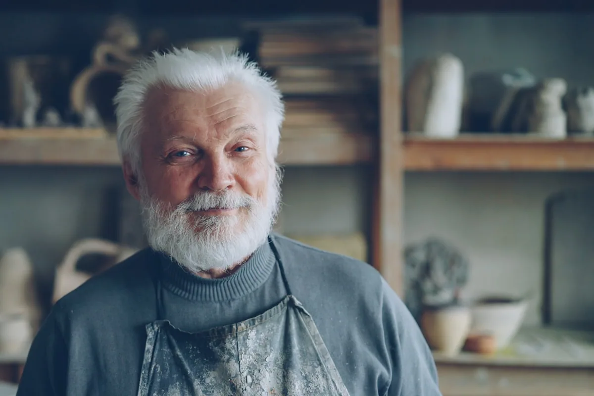 A grey-bearded maker in a workshop apron standing among shelves of finished pieces.