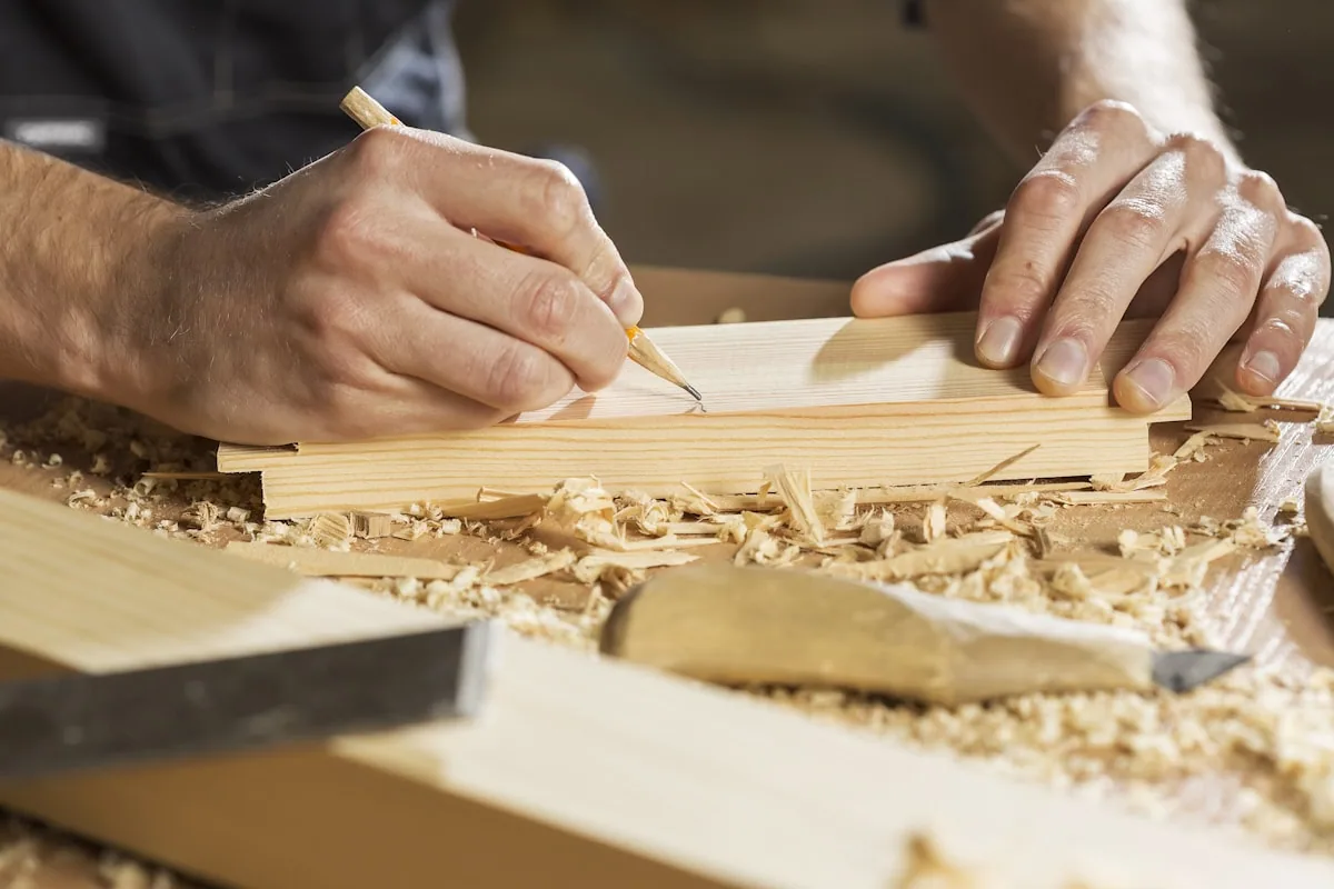 A joiner marking a length of pale timber with a pencil and square, shavings across the bench.